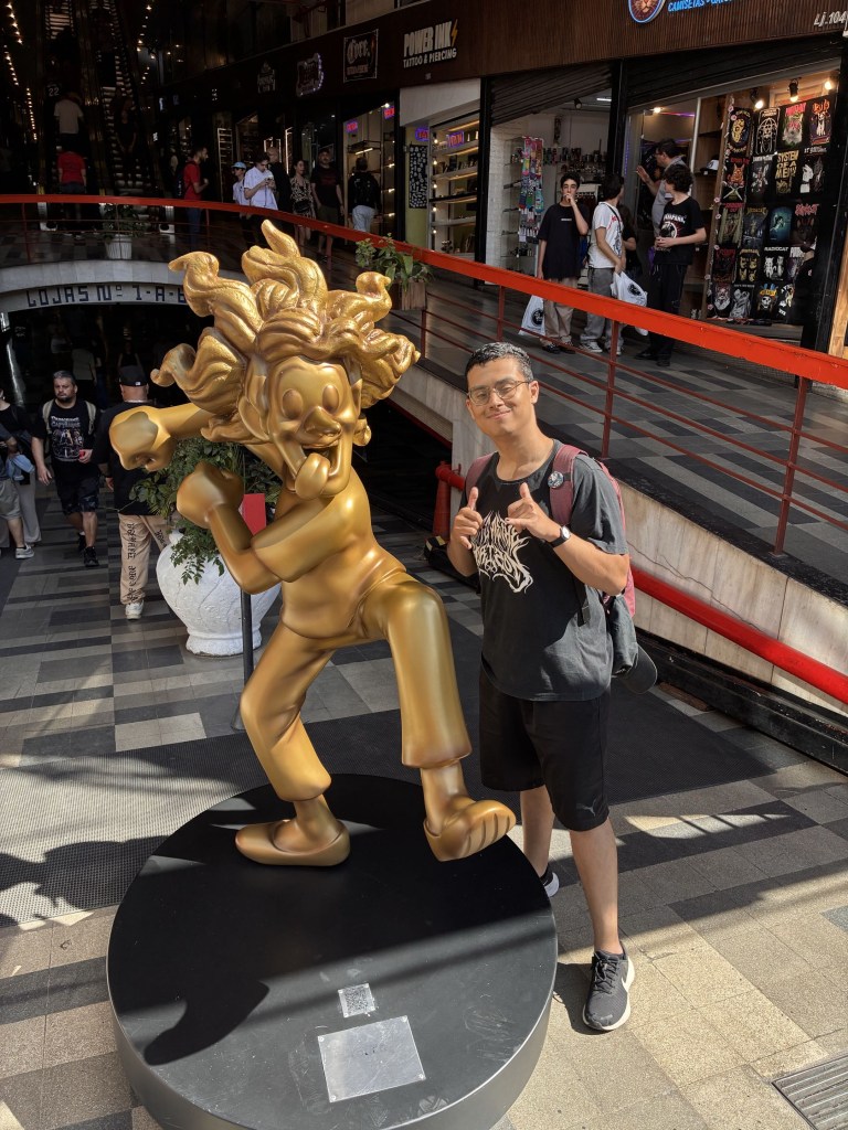 A young man standing on the right side of a gold statue indoors, smiling and making double "shaka" hand gestures. He has short dark curly hair, wears round glasses, a black t-shirt with a light-colored graphic, black shorts, dark sneakers, and carries a dark backpack. To his left stands a large golden cartoonish statue in a dynamic pose inside a brightly lit multi-level shopping arcade.