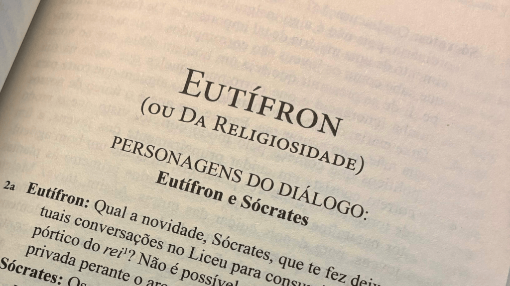 "Close-up photo of a page from a Portuguese edition of Plato's dialogue 'Eutífron' (Euthyphro), subtitled 'ou Da Religiosidade' (or On Religiosity). The page shows the heading 'PERSONAGENS DO DIÁLOGO: Eutífron e Sócrates' (Characters in the Dialogue: Euthyphro and Socrates), followed by the opening lines of the dialogue, with Eutífron asking Sócrates about his recent conversations at the King's Porch in the Lyceum."